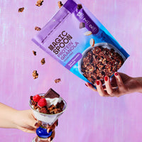 Hand holding Magic Spoon Dark Chocolate Peanut Butter granola box above yogurt parfait with berries and chocolate on pink bac