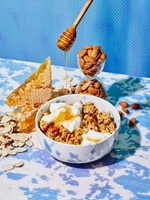 Honey almond granola in a white bowl with yogurt and honey drizzle, surrounded by almonds, honeycomb, and a honey dipper.