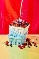 Four stacked pastel containers filled with granola, raspberries, and blueberries with milk pouring over top against red backg