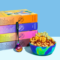 Four stacked colorful cereal boxes with a matching bowl filled with mixed cereal and a colorful spoon against a blue backgrou