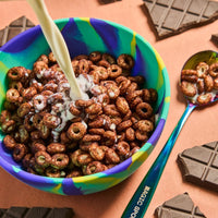 Chocolate cereal with milk being poured into a colorful bowl, with chocolate pieces and a spoon visible.